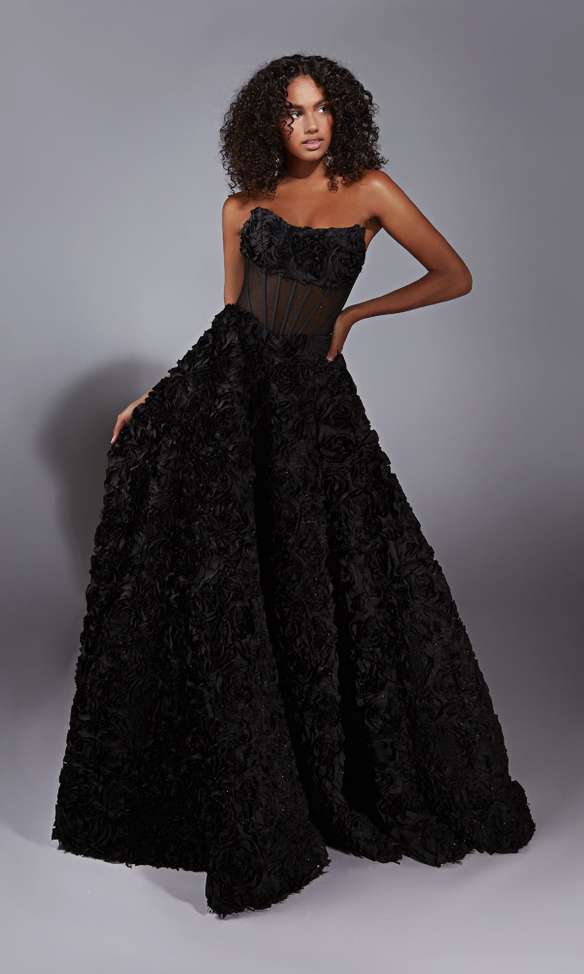A woman models the Alyce 61775 Strapless Rosette Prom Ball Gown, featuring a corset-style bodice and textured floral skirt, while standing against a plain gray background.