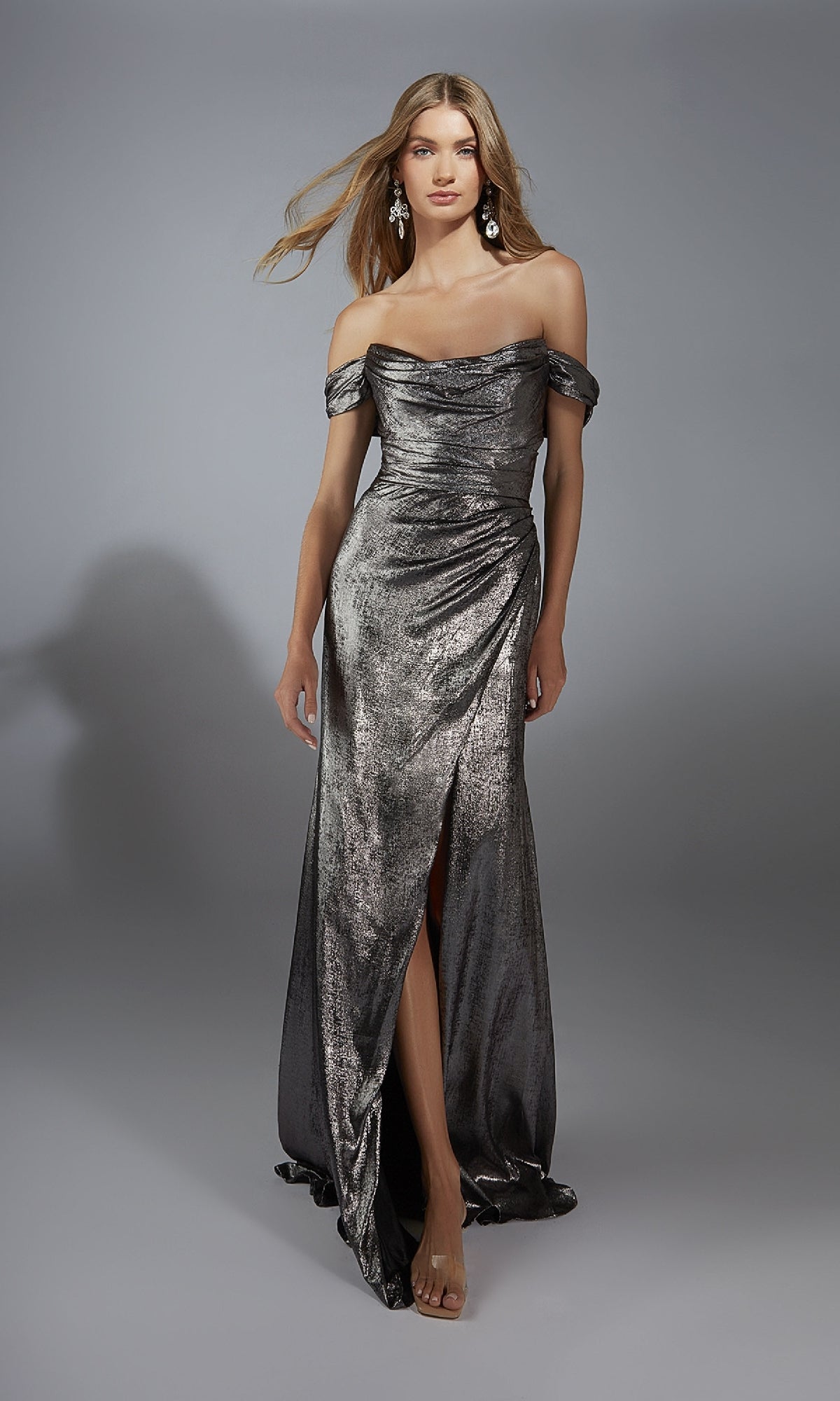 A woman stands against a plain gray background wearing the Alyce 61778 long prom dress, featuring a metallic off-the-shoulder design and a thigh-high slit.