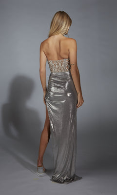 A woman faces away, wearing the Alyce 61780 long prom dress with a strapless beaded bodice, high slit, and shimmering metallic skirt against a plain gray background.