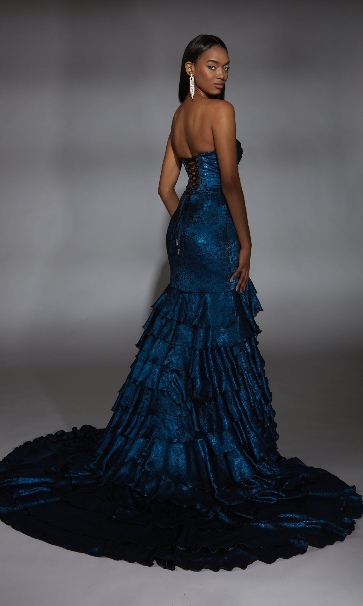 A woman models the Alyce 61782 long prom dress—strapless, dark blue mermaid style with ruffled layers and a sweeping train—posing elegantly against a plain gray background.