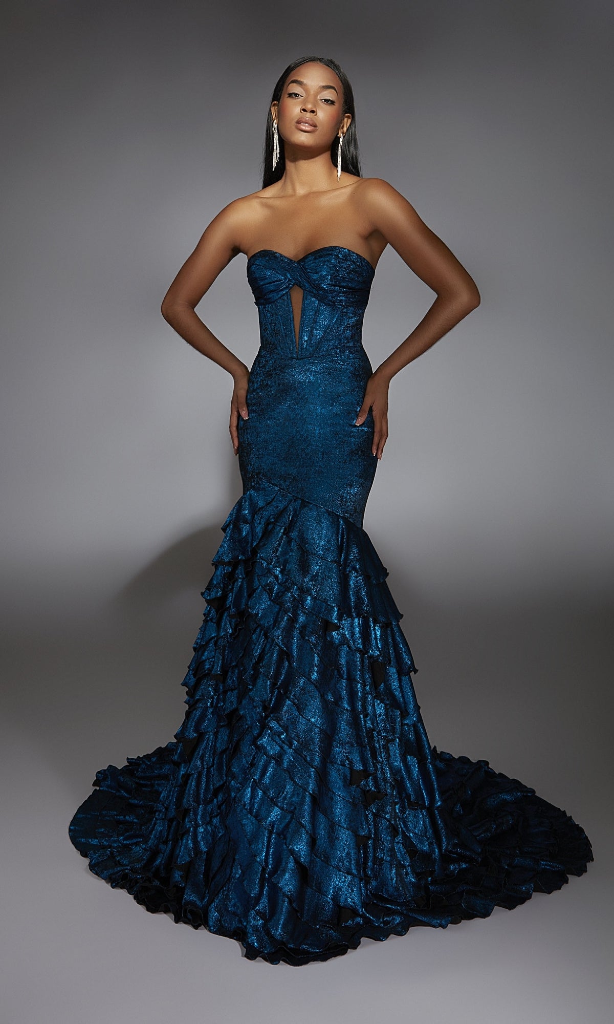 A woman models the Alyce 61782 long prom dress, a strapless dark blue mermaid gown with ruffled layers, a front cutout, and a modern fitted silhouette, standing against a plain gray background.