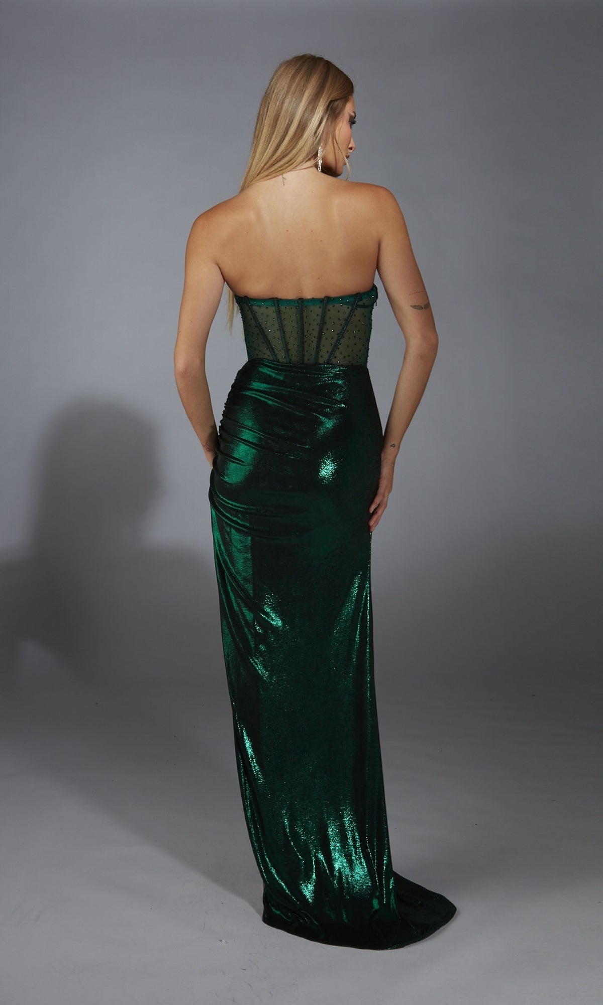 A woman faces away, modeling the Alyce 61784 long prom dress—a strapless metallic gown with a sheer corset bodice and floor-length dark green fabric—set against a plain gray background.
