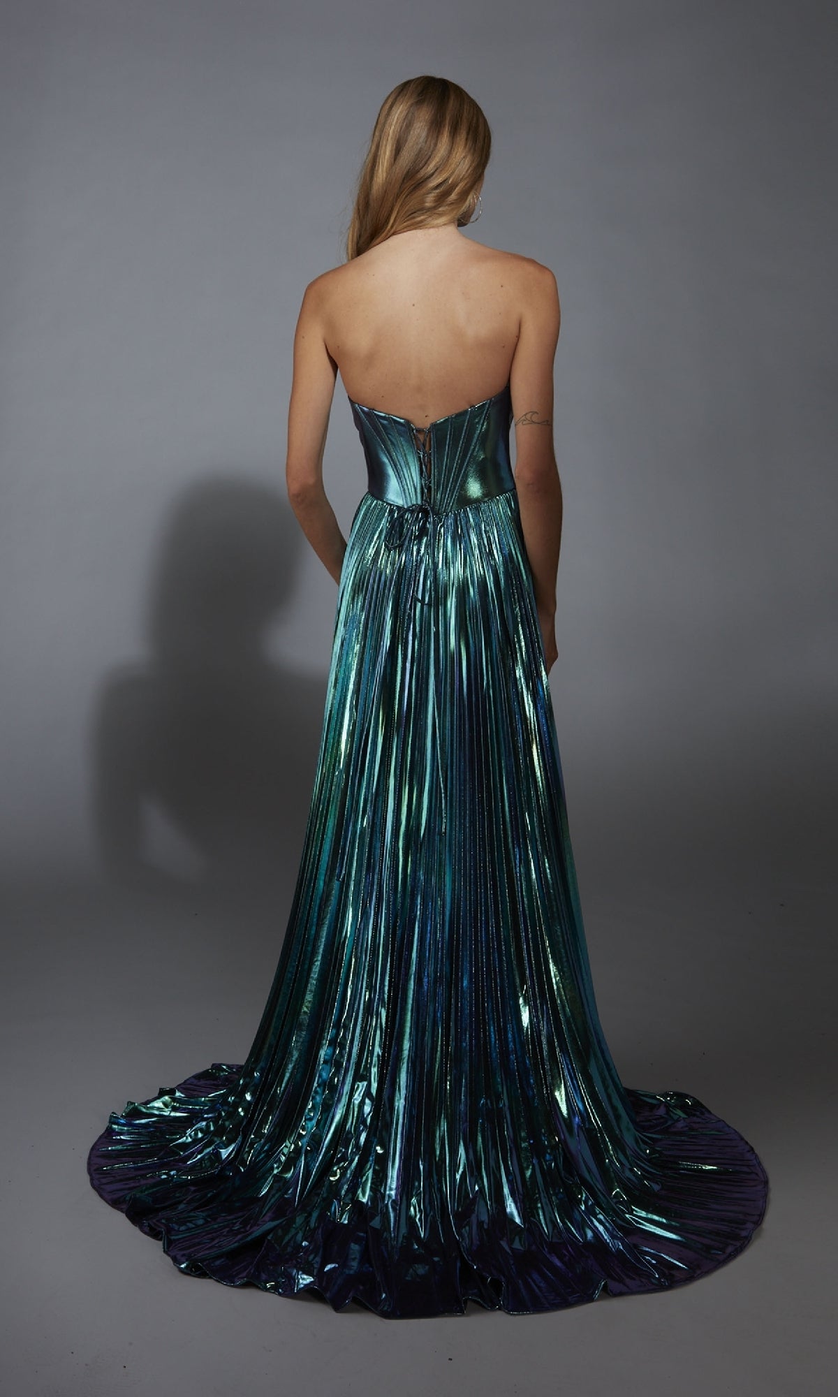 A woman with long blonde hair wears the Alyce 61786 Strapless Pleated Metallic Prom Gown, seen from the back against a gray background.