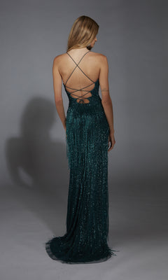 A woman with long hair stands facing away in the Alyce 61788, a sparkly dark teal prom dress with beaded fringe, a floor-length fringed skirt, and an open back with crisscross straps.