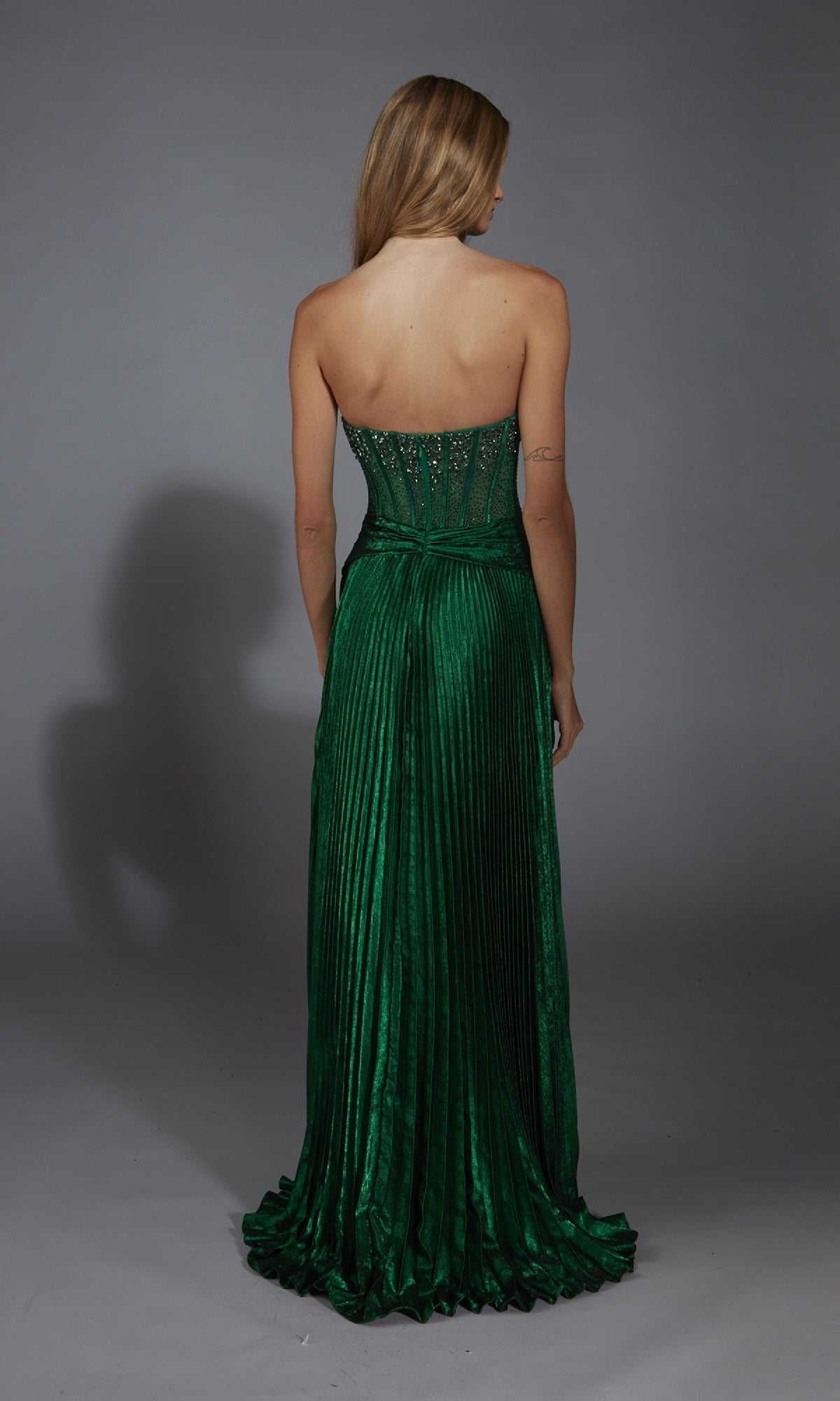 A woman faces away, wearing the Alyce 61789 Strapless Long Pleated Prom Dress in green with a fitted rhinestone corset bodice, set against a plain gray background.