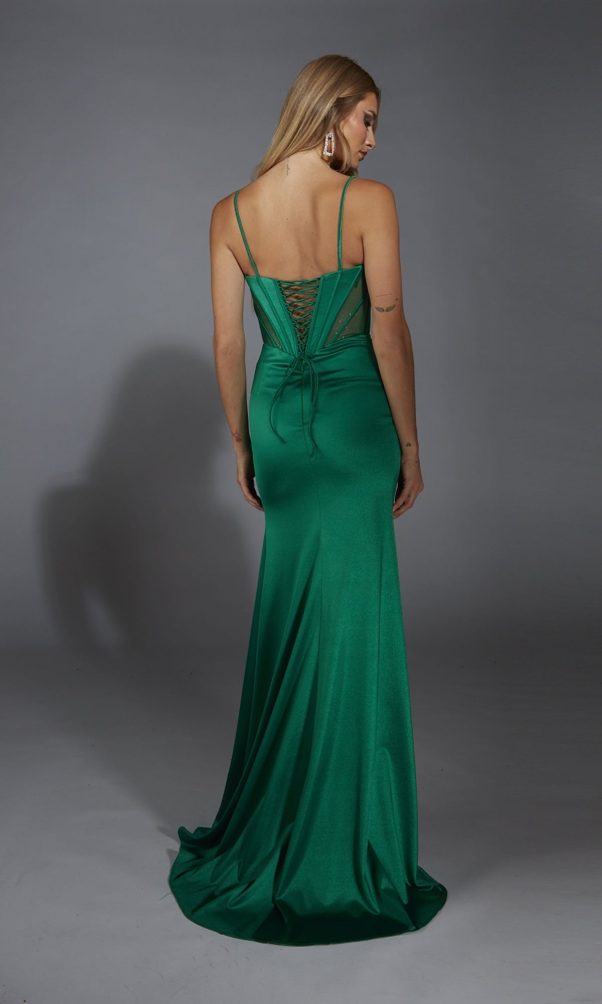 A woman faces away in the Alyce 61791 Lace-Up Long Corset Prom Dress, an emerald green, floor-length gown with thin straps and lace-up back detailing, set against a plain gray background.