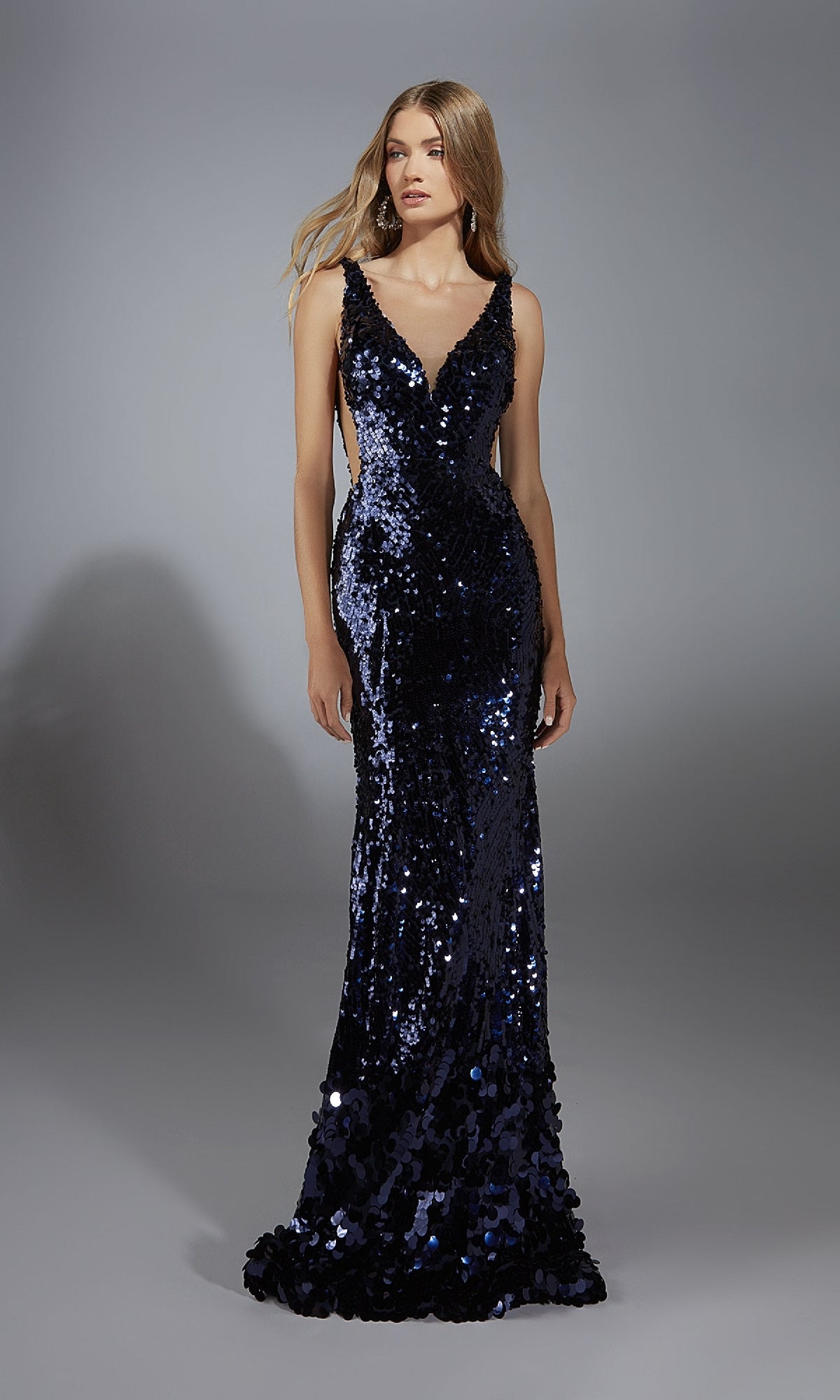 A woman wears the Alyce 61795 Sheer-Sides Long Sequin Prom Dress, a dark blue floor-length gown with a deep V-neckline, standing against a plain gray background.