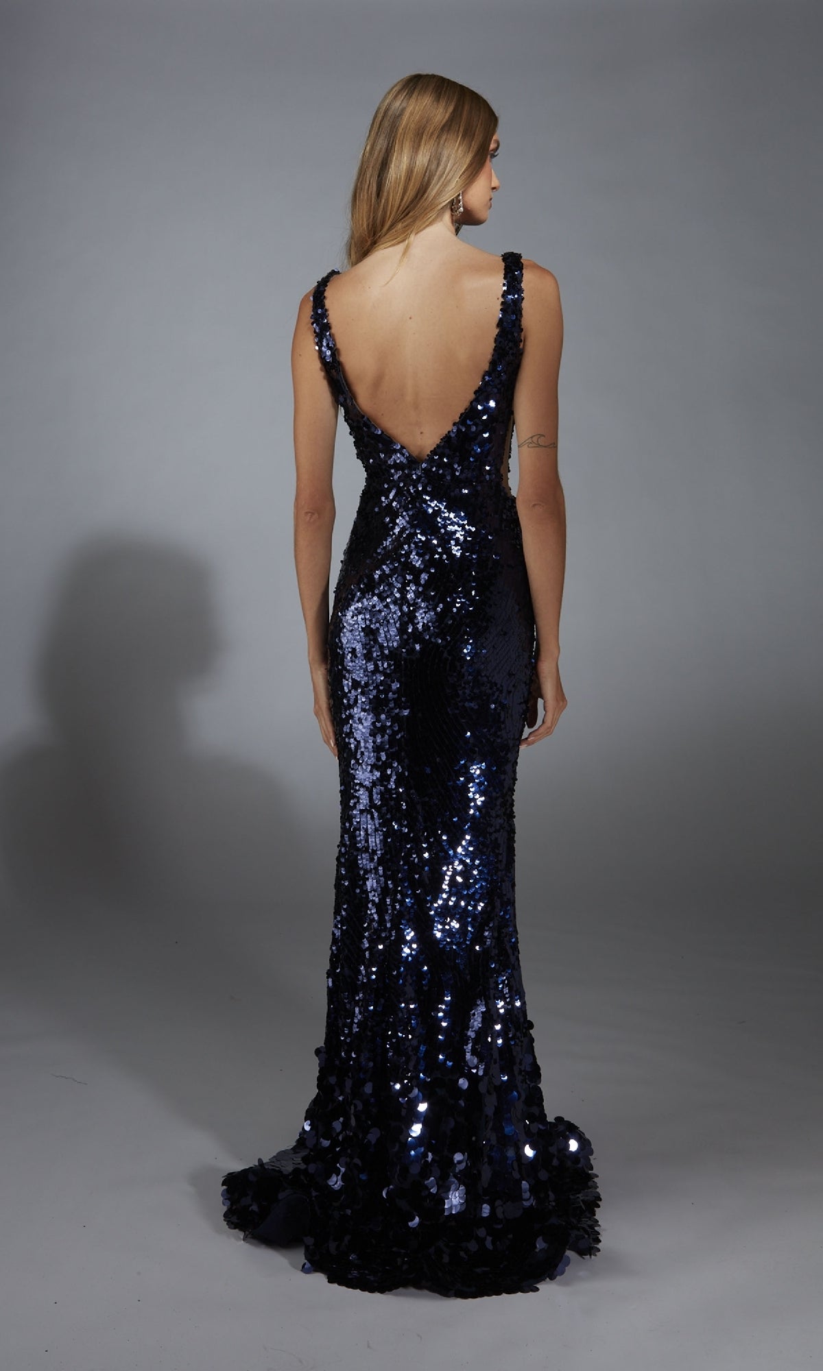 Woman in a dark blue Alyce 61795 Sheer-Sides long sequin prom dress with a deep V-back, standing on a gray backdrop and facing away from the camera.