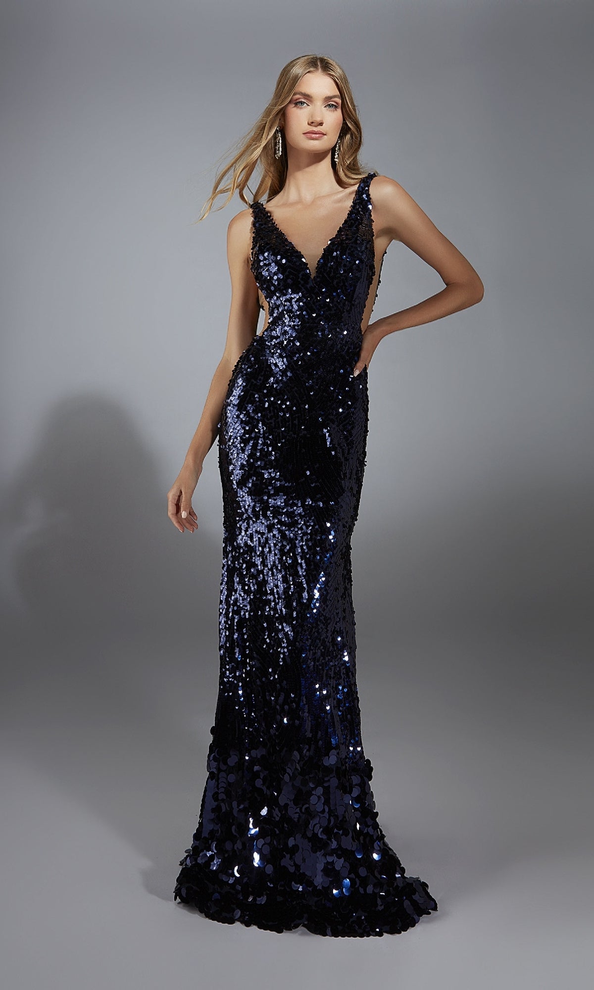 A woman poses against a gray background, wearing the Alyce 61795 Sheer-Sides Long Sequin Prom Dress—a sleeveless, floor-length dark blue gown with a deep V-neckline.