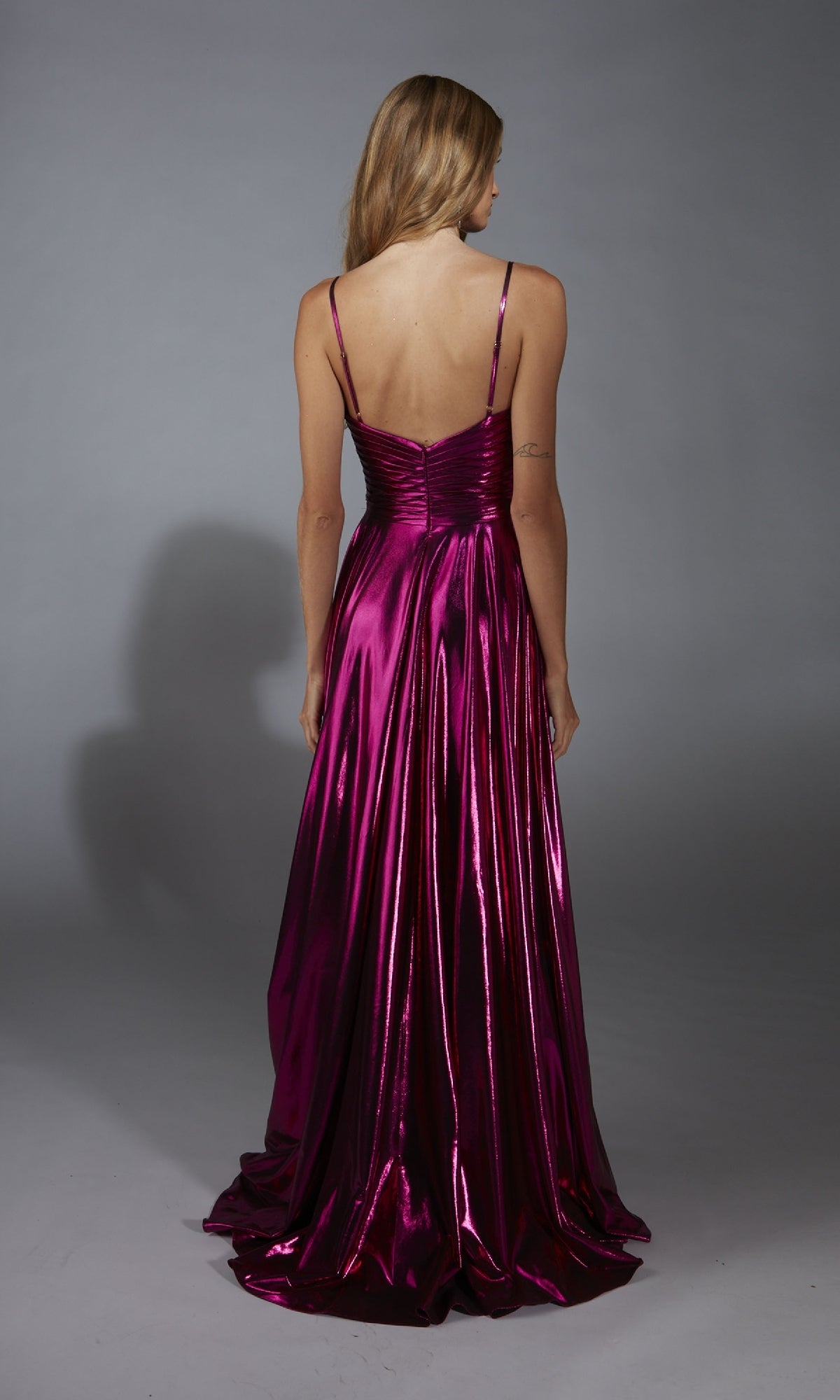 A woman faces away, wearing the Alyce 61801 long metallic A-line prom dress with spaghetti straps. The floor-length gown shines in magenta against a plain gray background.