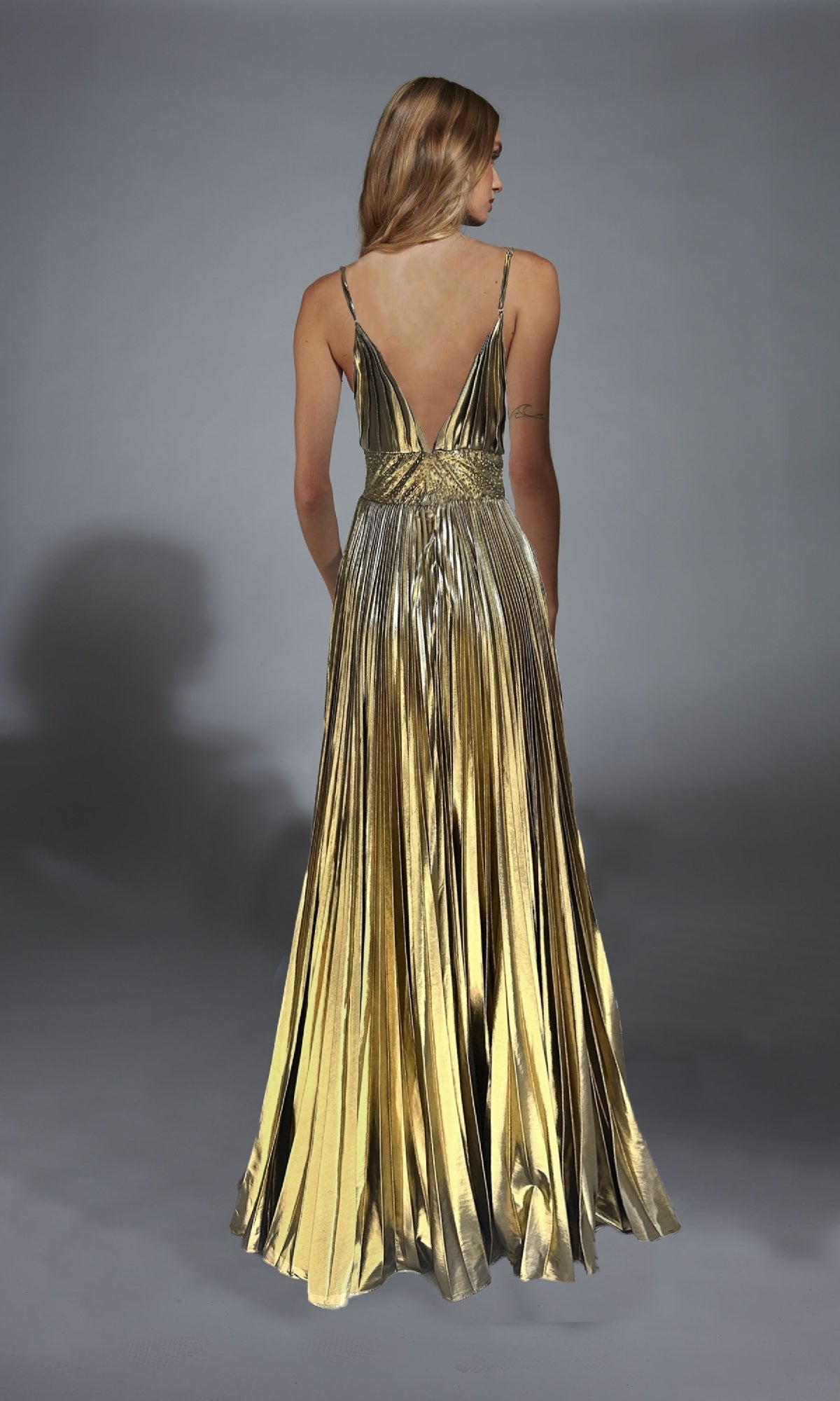 A woman faces away in the Alyce 61804 Long Prom Dress, showcasing its metallic gold finish, pleated A-line skirt, and flowing floor-length design against a neutral backdrop.