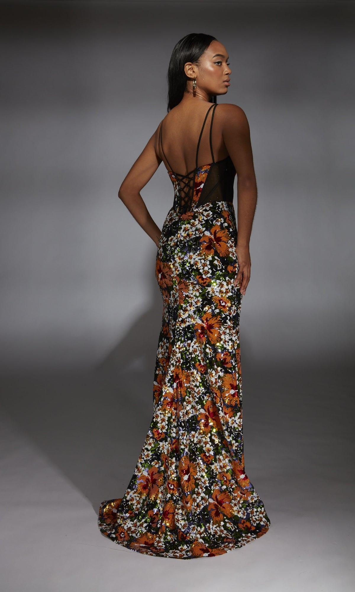 A woman wears the Alyce 61809 long prom dress with floral sequin accents, black sheer back, and thin straps, standing in a studio facing away.