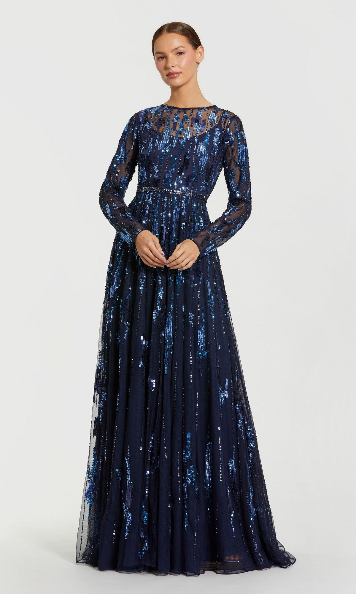 A woman wearing the Mac Duggal 6181 Long Formal Dress in Midnight, featuring a fitted bodice with sheer long sleeves and sequin detailing, stands against a plain light background.