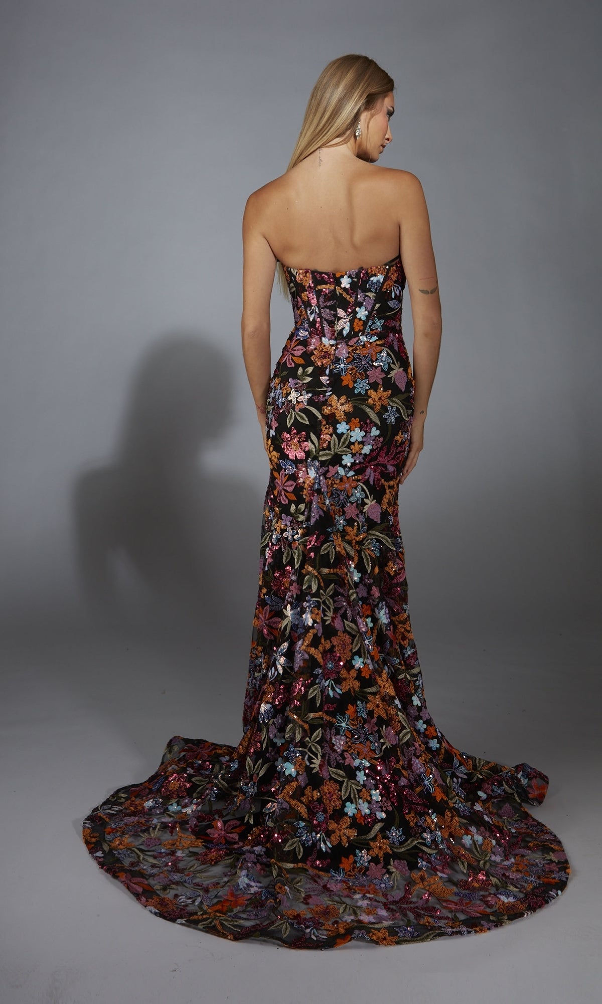 A woman faces away, wearing the Alyce 61810 long prom dress—a strapless mermaid gown with a sequin flower print and long train—against a neutral background.