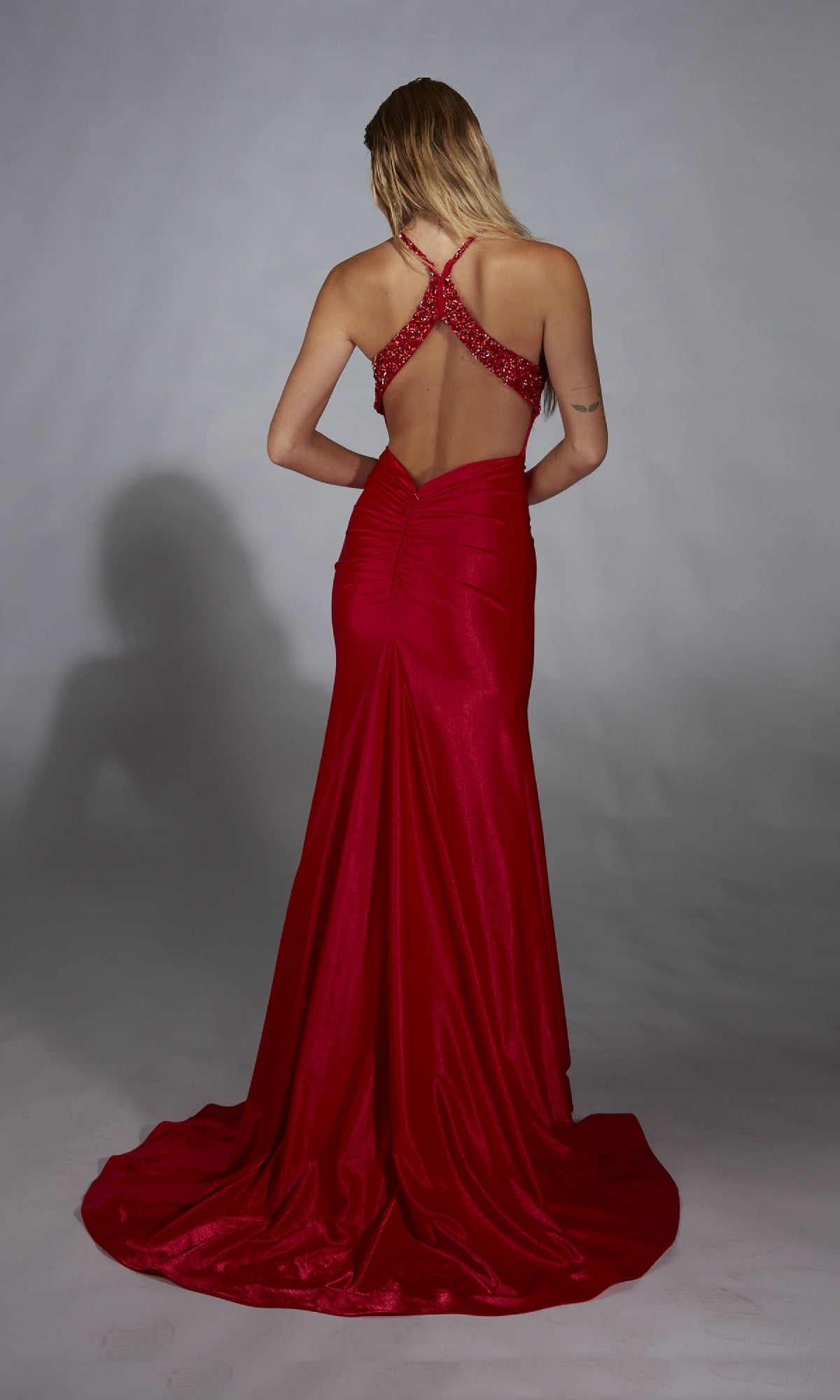 A woman stands facing away, wearing the Alyce 61820 long prom dress in red with shimmery jersey fabric, crisscross straps, an open back, and center ruching.