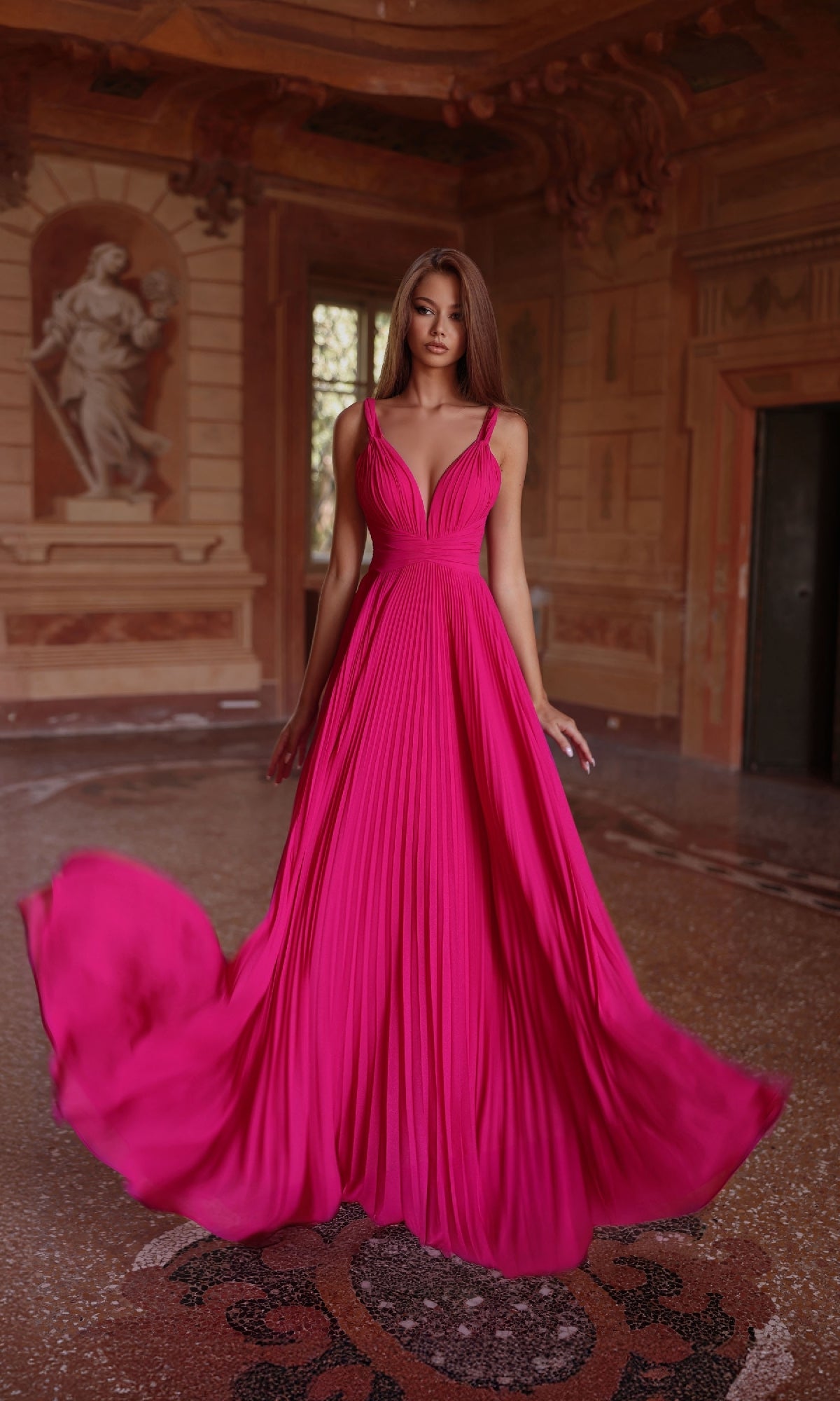 A woman stands in an ornate room wearing the Alyce 61823, a vibrant fuchsia long prom dress with a V-neckline and flowing A-line skirt, her hair down as the pleated fabric billows outward.