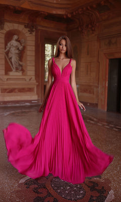A woman stands in an ornate room wearing the Alyce 61823, a vibrant fuchsia long prom dress with a V-neckline and flowing A-line skirt, her hair down as the pleated fabric billows outward.