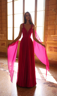 A woman stands indoors in the Alyce 61823—a floor-length, deep pink prom dress featuring an a-line skirt, pleated details, and sheer side panels—while sunlight streams through large windows behind her.