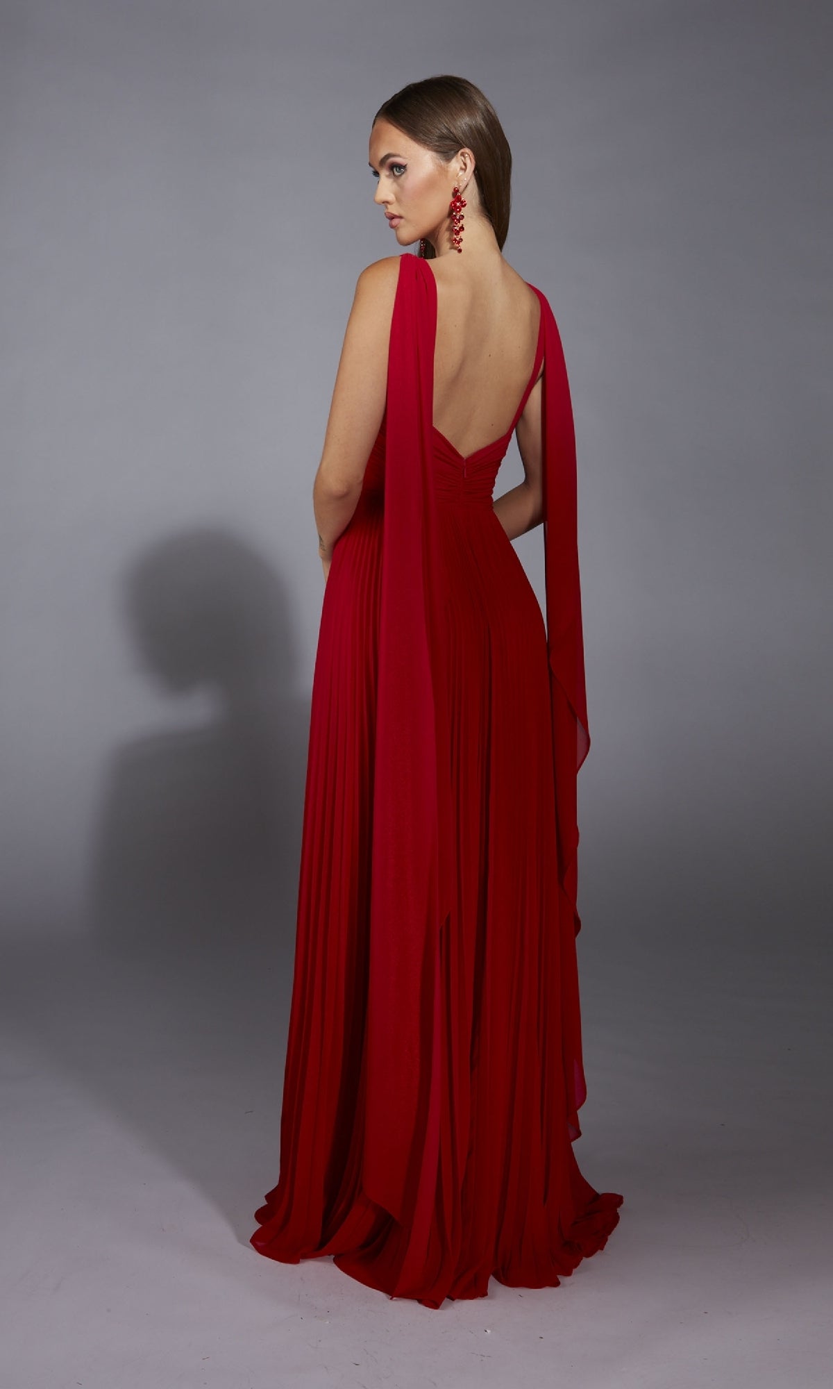 A woman wearing the Alyce 61823 long prom dress with a v-neckline and draped red fabric, paired with matching red earrings, stands facing away against a plain gray background.