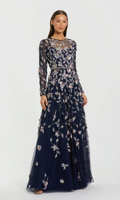 A woman models the Mac Duggal 6183 Long Sleeve Beaded Formal Gown, featuring navy blue fabric, sheer long sleeves, and floral embroidery, standing against a plain white background.