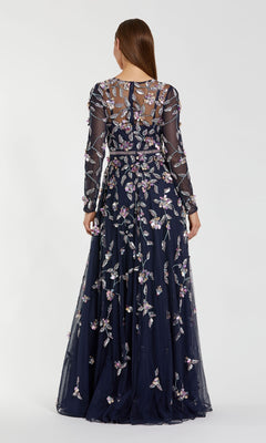 A woman stands facing away in the Mac Duggal 6183 Long Sleeve Beaded Formal Gown, a floor-length navy blue dress featuring sheer fabric and intricate floral embroidery.