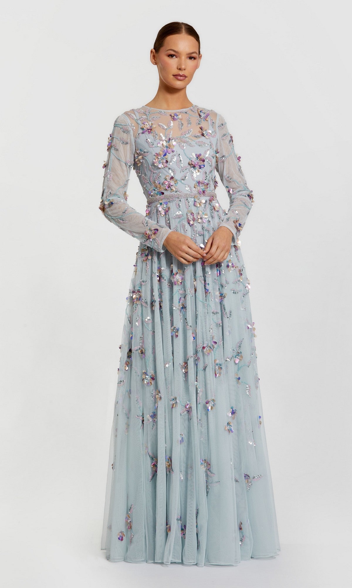 A woman wears the Mac Duggal 6183, a pale blue floor-length formal gown with long sleeves, beadwork, sheer panels, and colorful floral embroidery, standing against a plain light background.