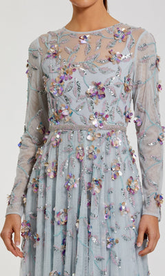 A woman wearing the Mac Duggal 6183 Long Sleeve Beaded Formal Gown—an elegant light blue sheer dress featuring embroidered floral patterns and 3D sequin flower embellishments.