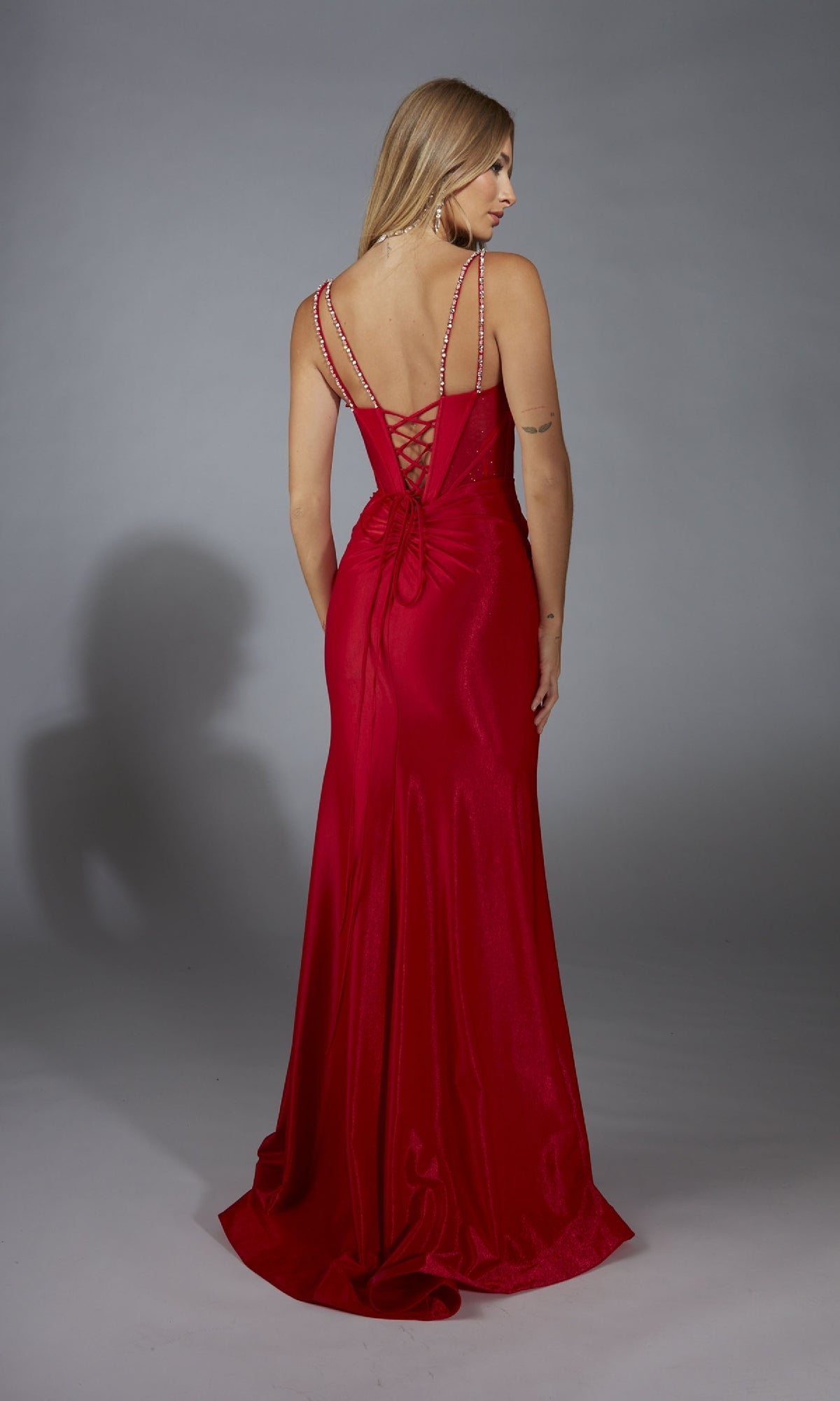 A woman faces away, wearing the Alyce 61830 long prom dress with a fitted silhouette, lace-up corset bodice, and thin beaded straps, set against a plain gray background.