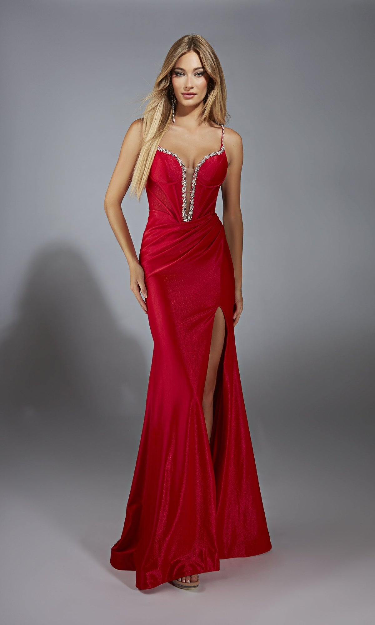 A woman models the Alyce 61830 Long Prom Dress—a sleeveless, floor-length red gown with a deep neckline, lace-up corset bodice, embellished straps, and high slit—against a plain gray background.