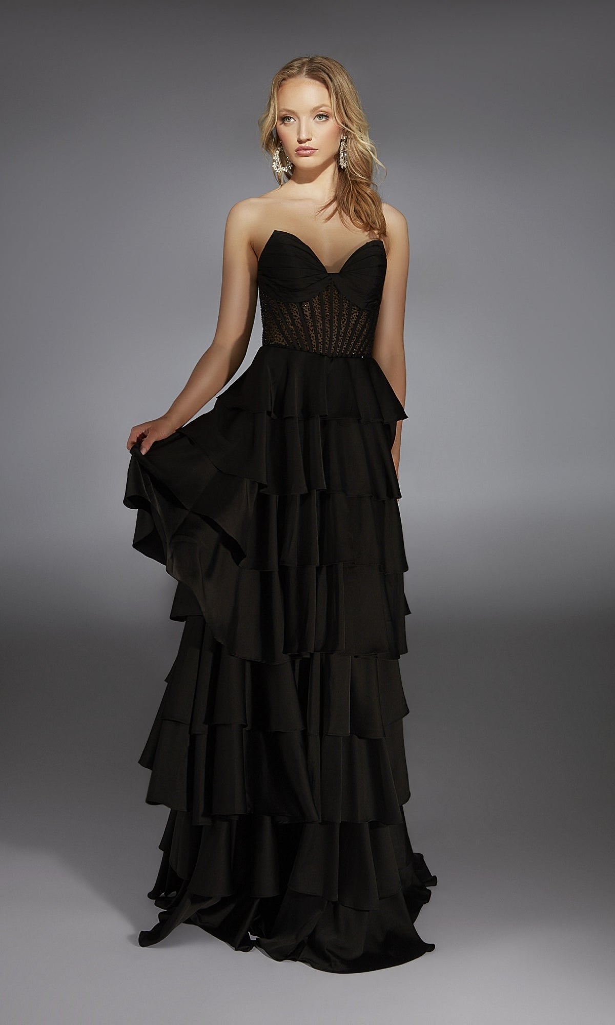 A woman in the Alyce 61833 long prom dress, featuring a strapless black design with a fitted sheer corset bodice and ruffle tiered A-line skirt, stands against a neutral background.