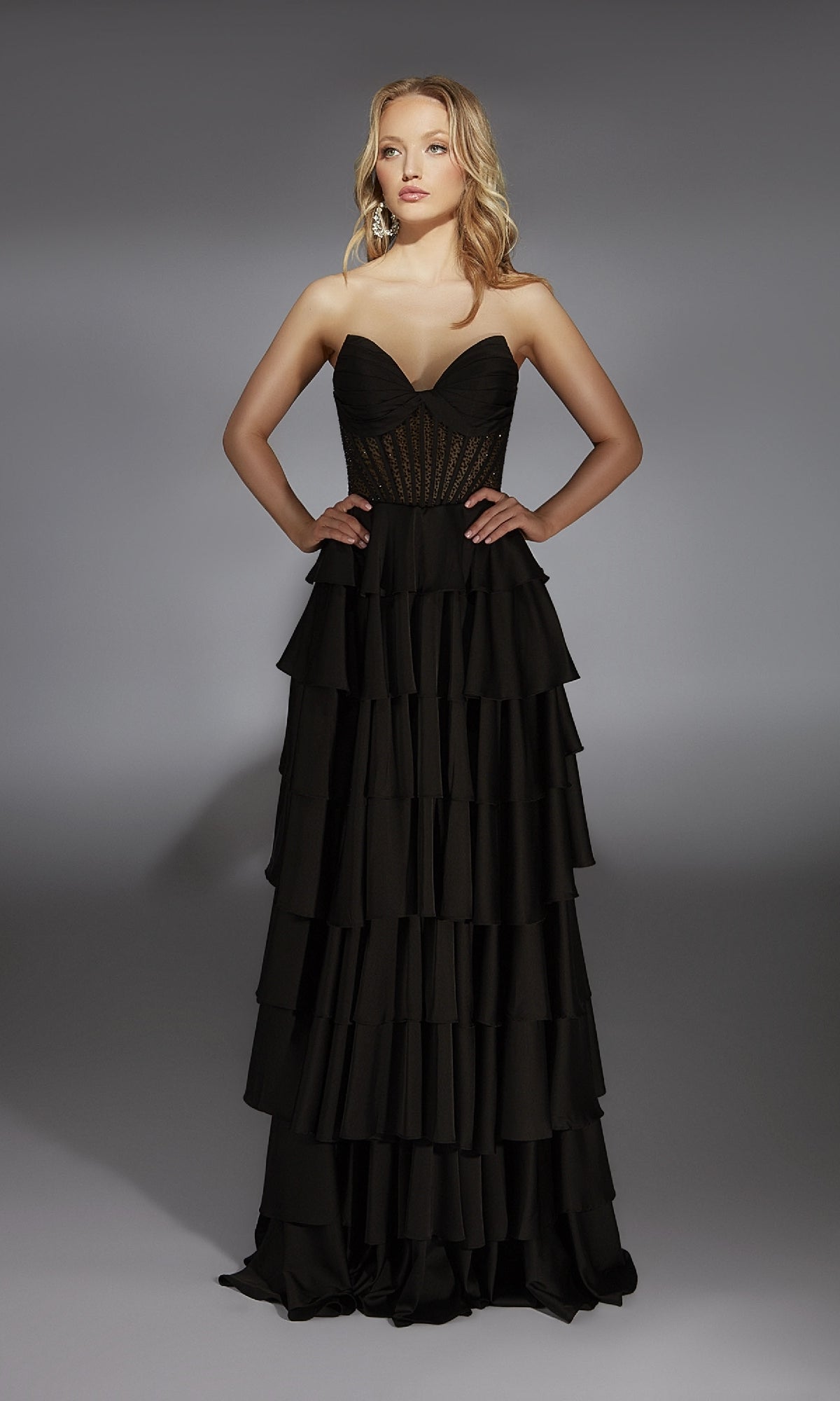 A woman stands with hands on hips, wearing the Long Prom Dress: Alyce 61833—a strapless gown with a fitted sheer corset bodice and dramatic ruffle tiered a-line skirt—against a plain gray background.