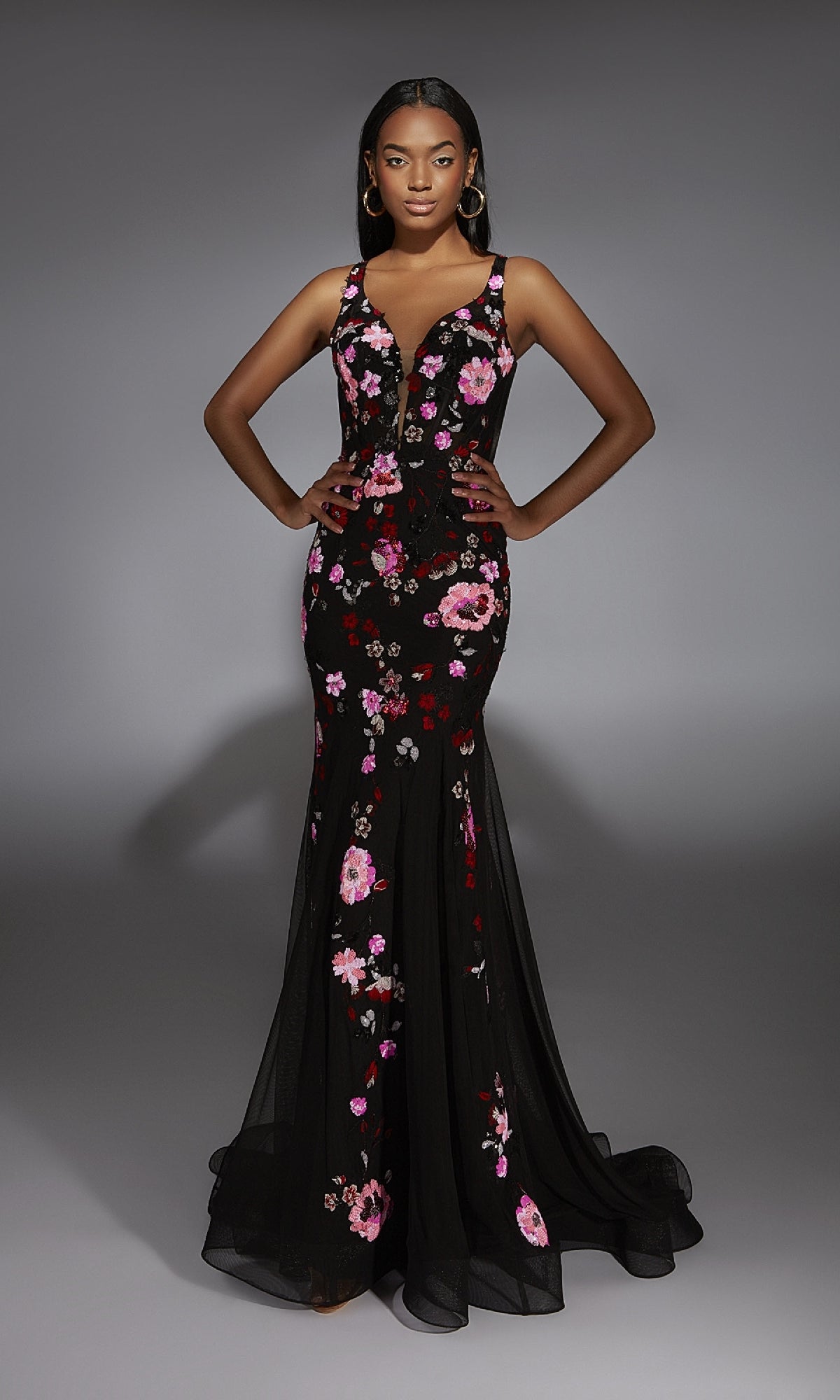 A woman stands with hands on hips, wearing the Long Prom Dress: Alyce 61839—a fitted black mermaid dress with pink and red floral details—against a plain gray background.