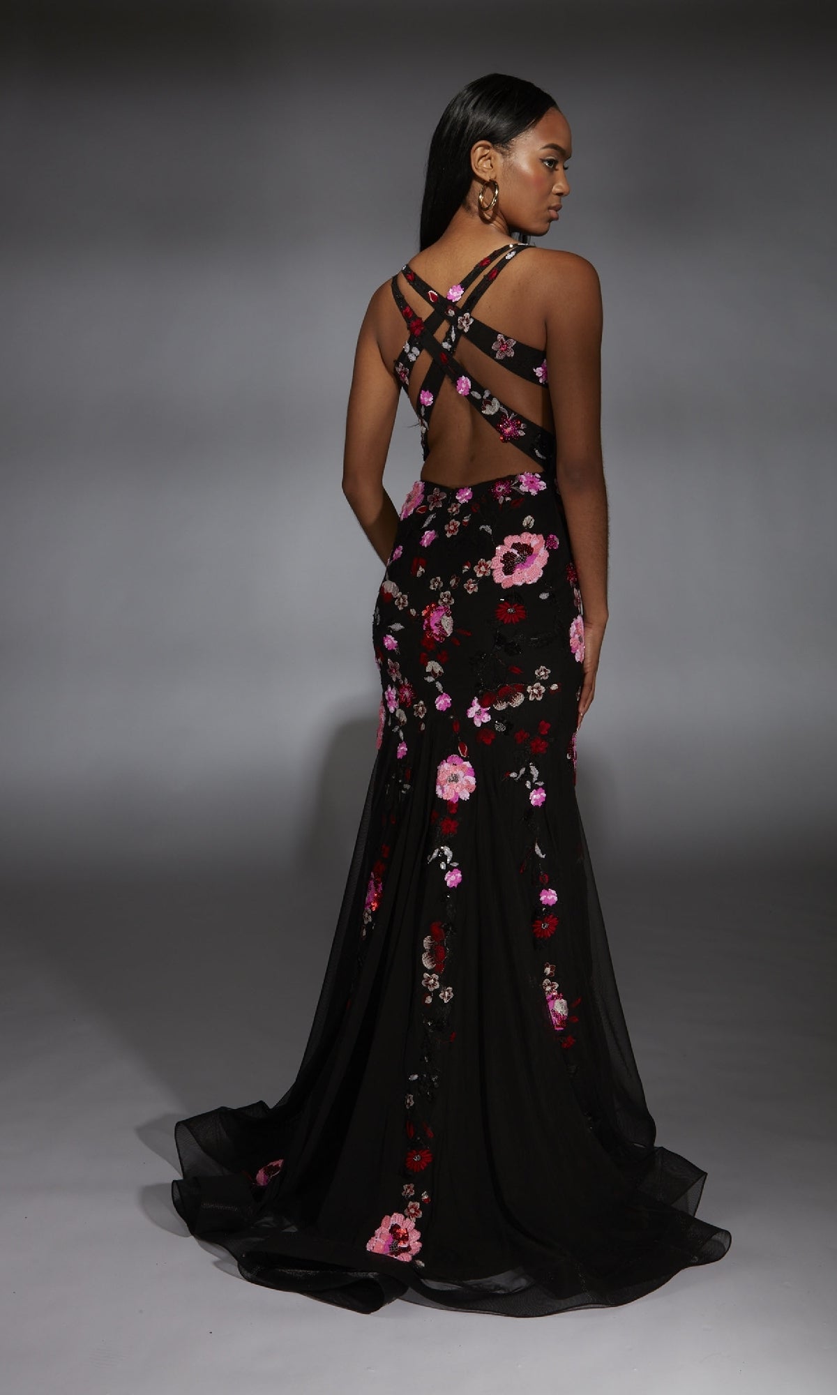 A woman wearing the Alyce 61839 Long Prom Dress, a black floral mermaid gown with sequin details and crisscross back straps, stands on a neutral background.