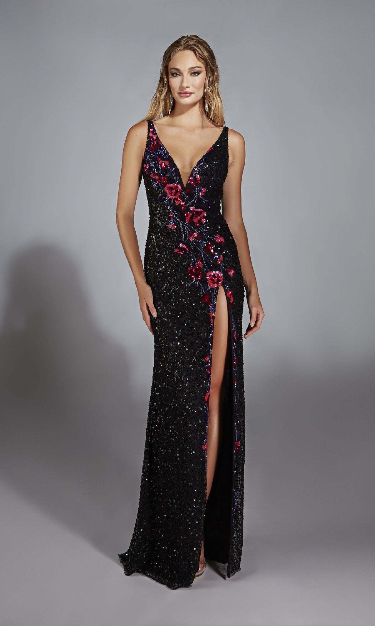 A woman models the Alyce 61841 long prom dress, a sleeveless black sequined gown with a deep V-neck, bold red floral pattern, and high slit, set against a plain gray background.