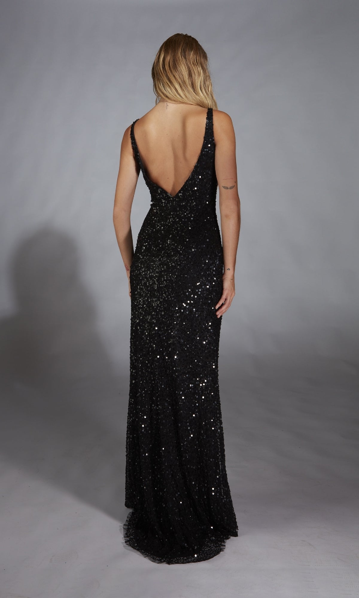 A person in the Alyce 61841 long black prom dress, featuring a floor-length sleeveless design and deep V-back, stands facing away from the camera against a gray background.