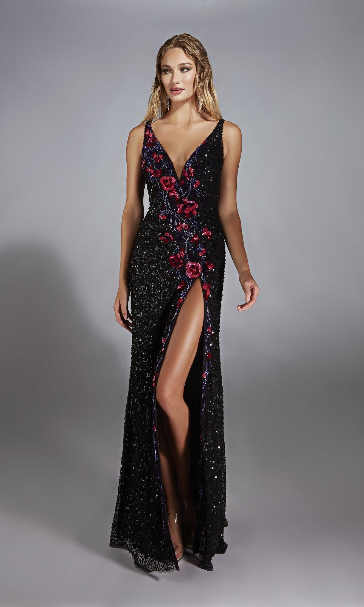 A woman stands against a plain gray background wearing the Alyce 61841 long prom dress—a sleeveless black sequin gown with a deep V-neck, floral embroidery, and a high slit.