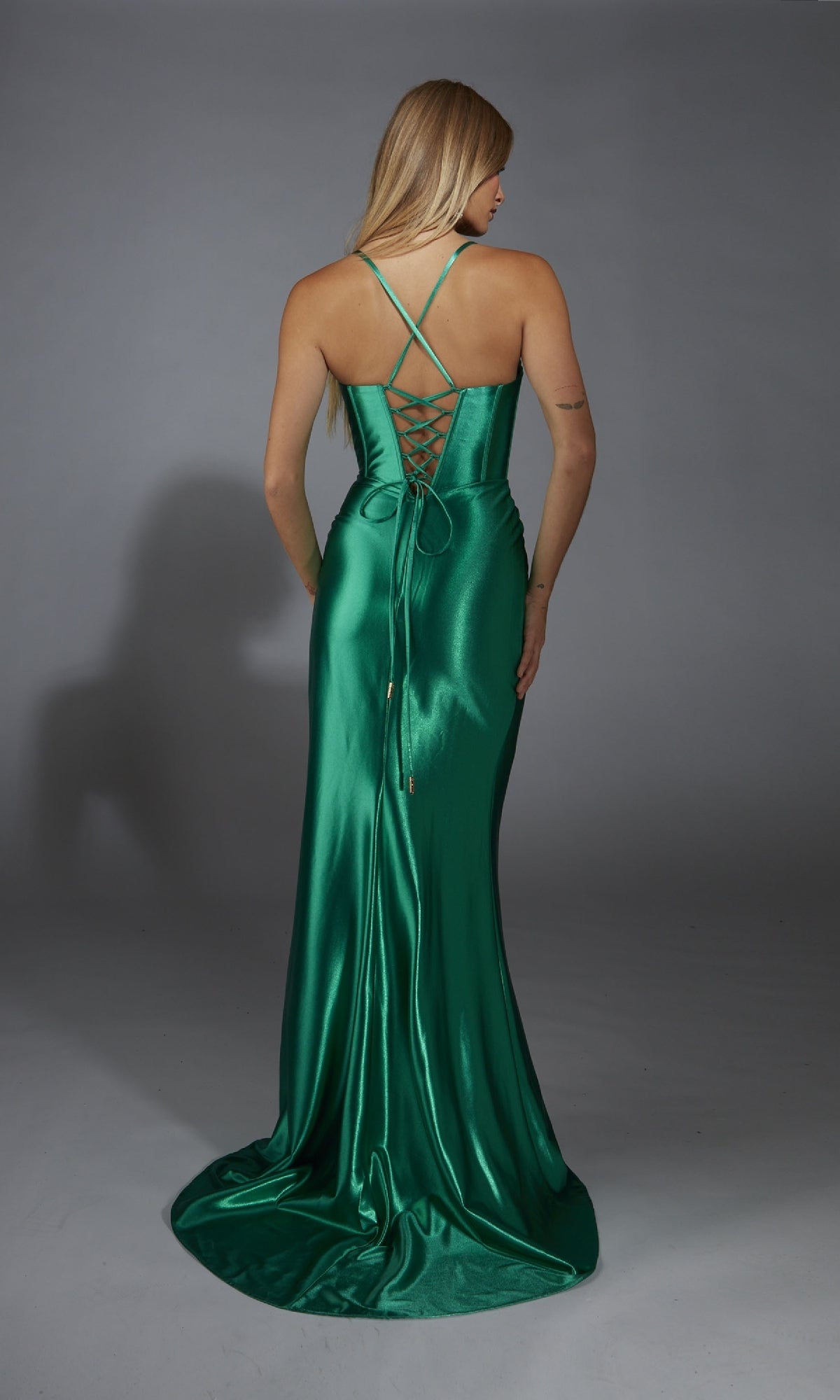 A woman stands facing away from the camera in a shiny green Alyce 61849 long prom dress featuring metallic stretch satin, spaghetti straps, and a lace-up back against a plain background.