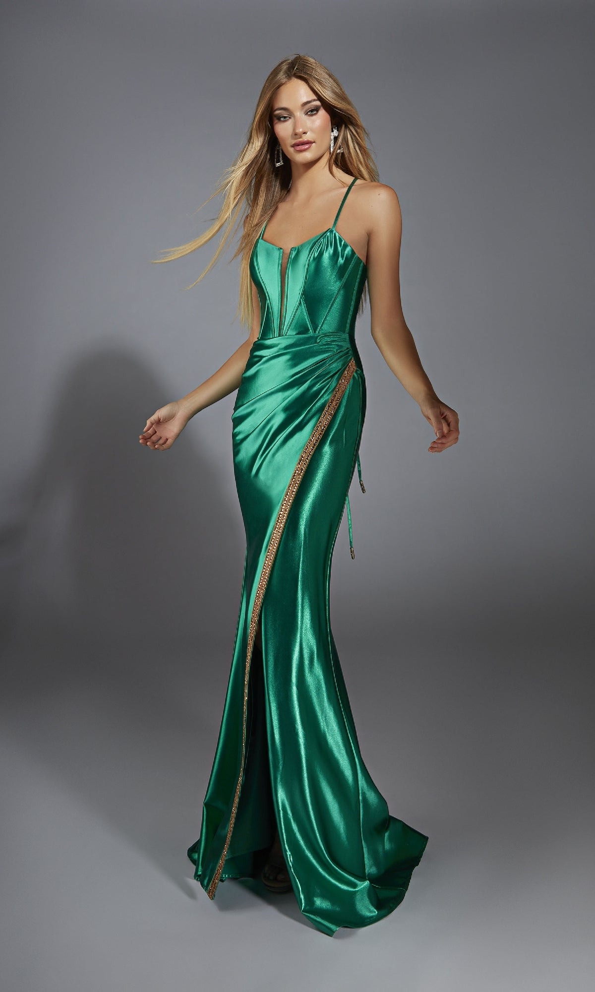 A woman poses against a plain gray backdrop wearing the Alyce 61849 long prom dress in emerald green metallic stretch satin, featuring a fitted silhouette, thin straps, corset bodice, and gold detailing.