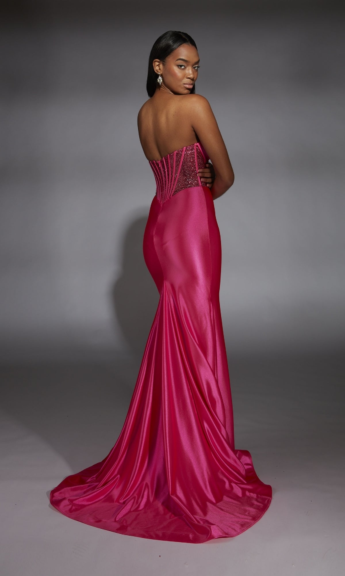 A woman poses with her back to the camera against a gray background, wearing the Alyce 61851: a strapless, fitted hot pink long corset prom dress with a flared train.