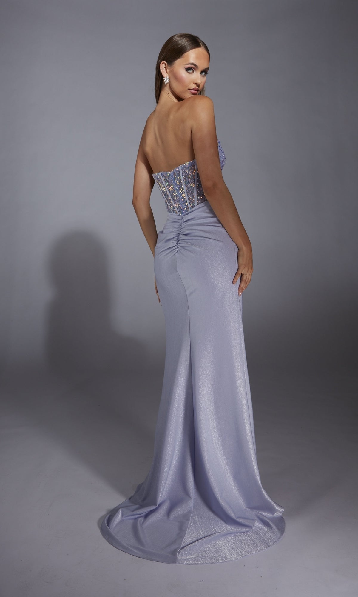 A woman wearing the Alyce 61856 long prom dress—strapless, metallic light purple with an embellished corset bodice and floor-length skirt—stands looking over her shoulder against a plain gray background.