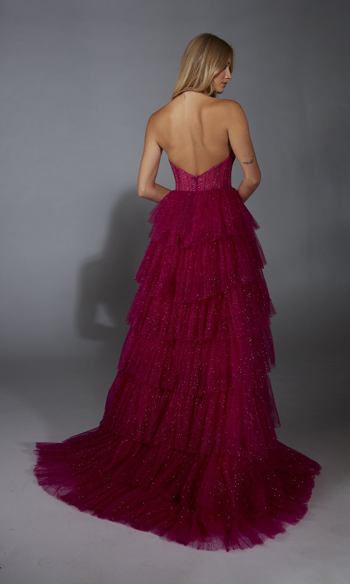 A woman stands with her back to the camera against a plain grey background, wearing the Alyce 61859 long prom dress—a strapless, backless magenta tiered gown with a corset bodice.