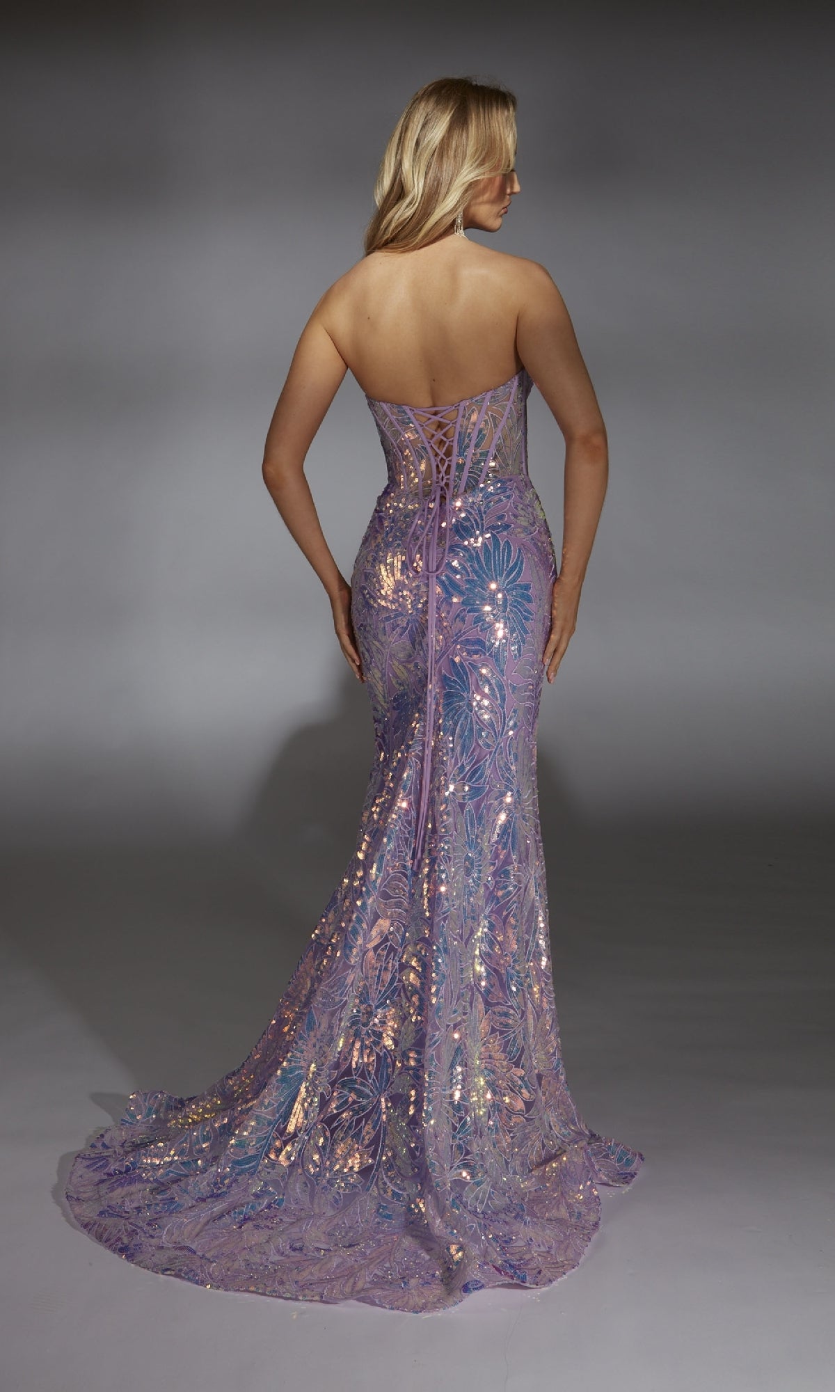 A woman faces away, wearing the Alyce 61862 long prom dress—a strapless, form-fitting lilac mermaid gown with sequins, a lace-up back, and a floor-length lavender design set against a plain gray background.