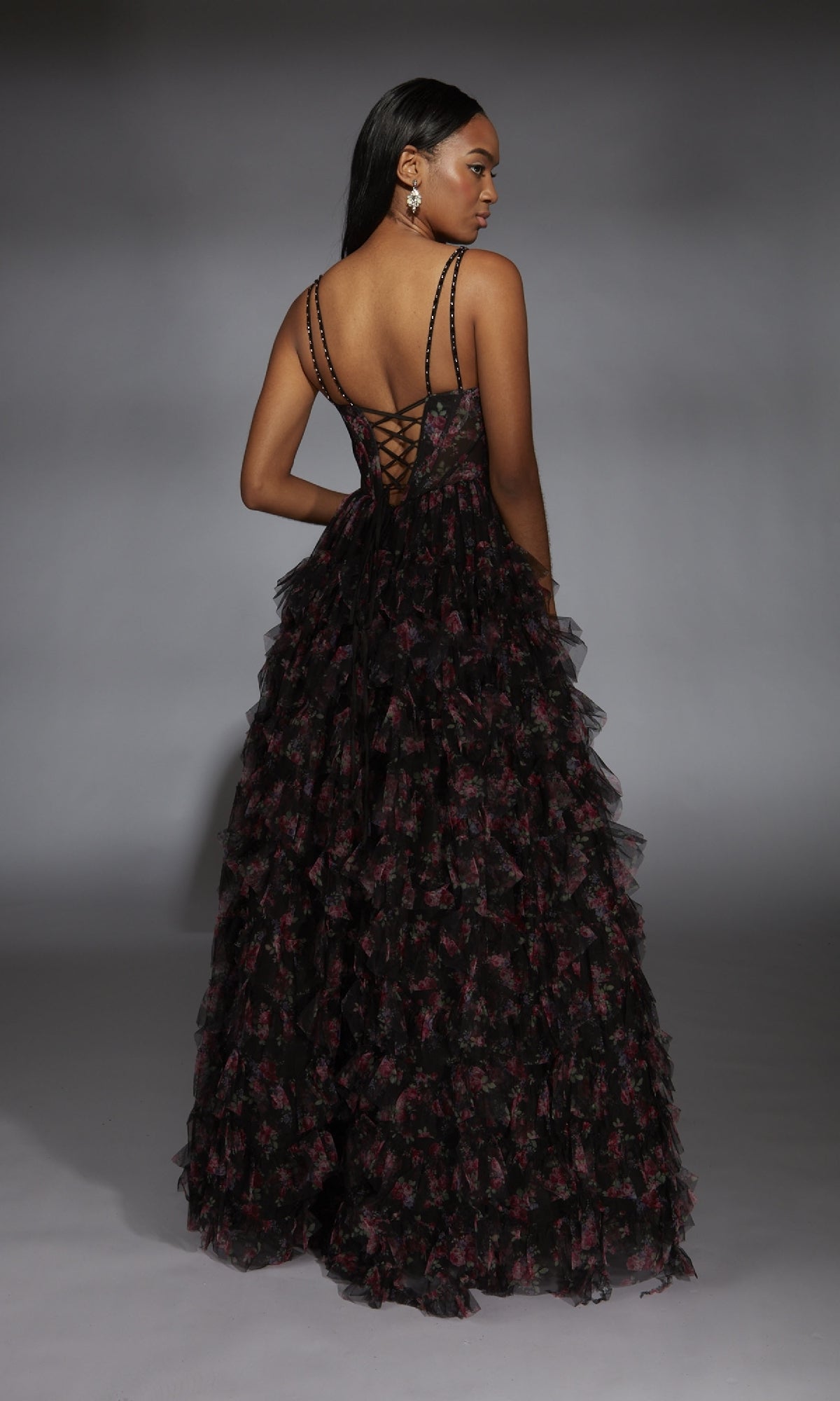 A woman stands sideways, looking over her shoulder in a black Alyce 61866 long prom dress featuring a floor-length ruffle tiered A-line skirt and lace-up corset bodice, set against a gray studio backdrop.