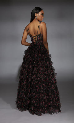 A woman stands sideways, looking over her shoulder in a black Alyce 61866 long prom dress featuring a floor-length ruffle tiered A-line skirt and lace-up corset bodice, set against a gray studio backdrop.