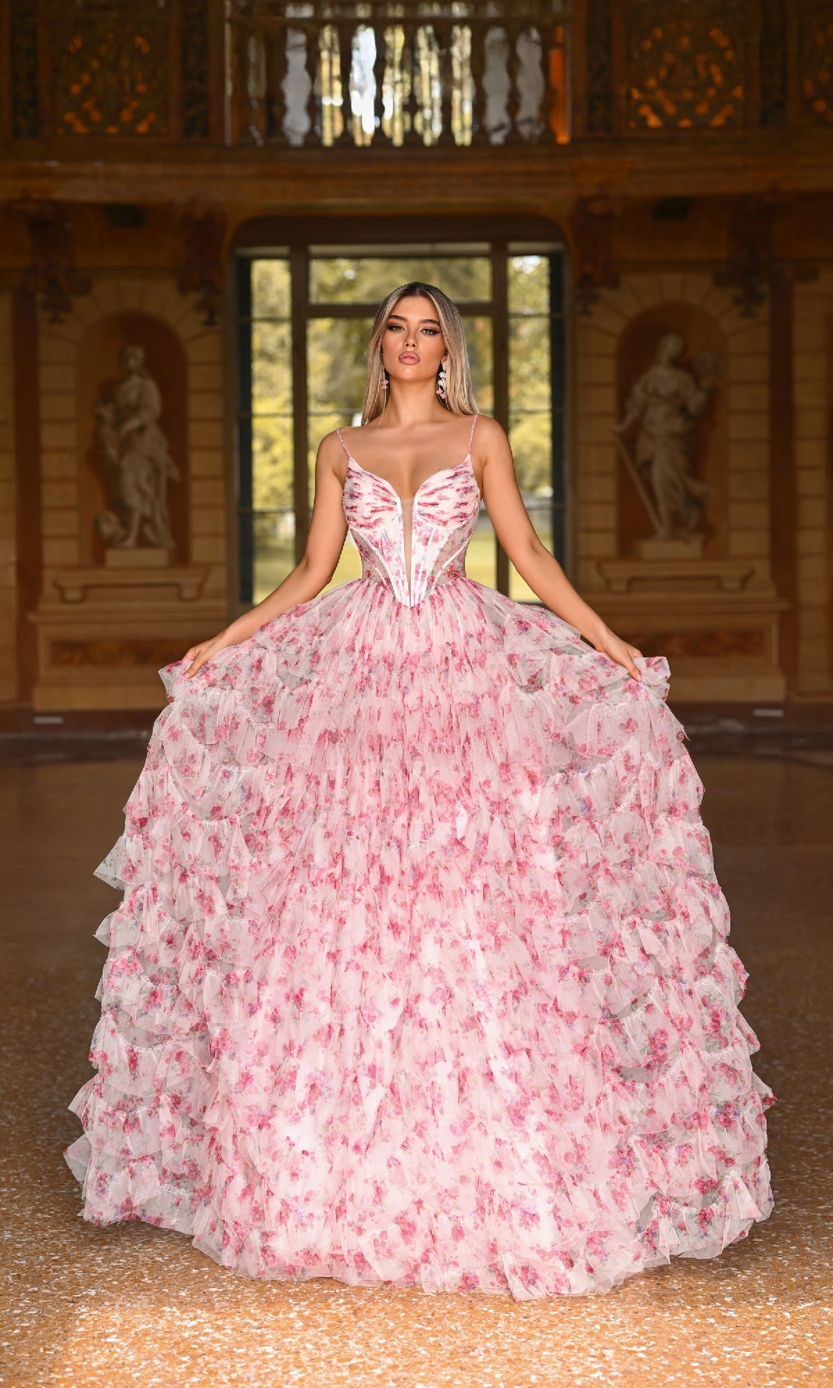 Indoors, a woman displays the Alyce 61866 long prom dress—voluminous and pink with a printed design, corset bodice, and deep neckline; statues and big windows are visible in the background.