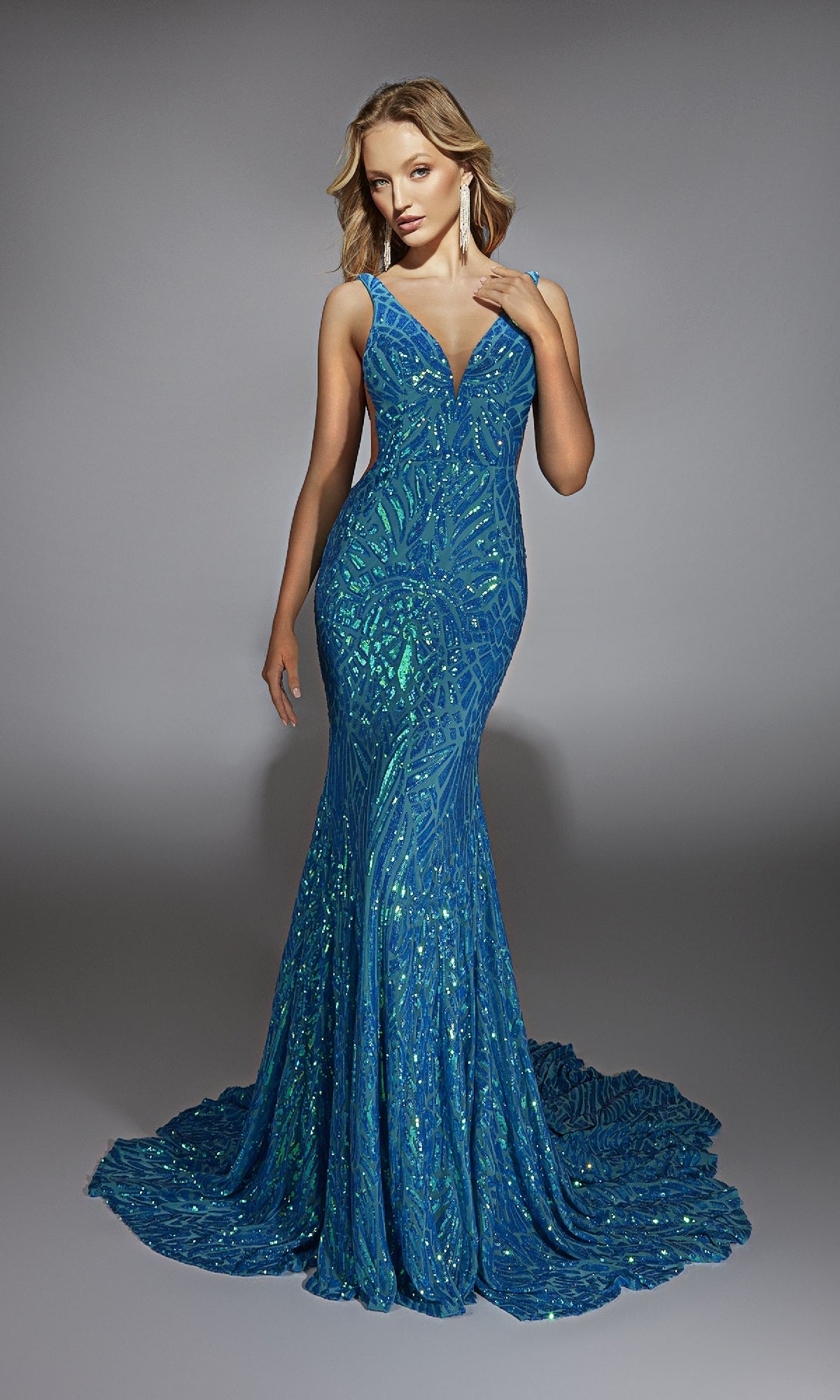 A woman stands against a plain gray background wearing the Alyce 61876 long prom dress, a form-fitting blue gown adorned with sparkling sequins.