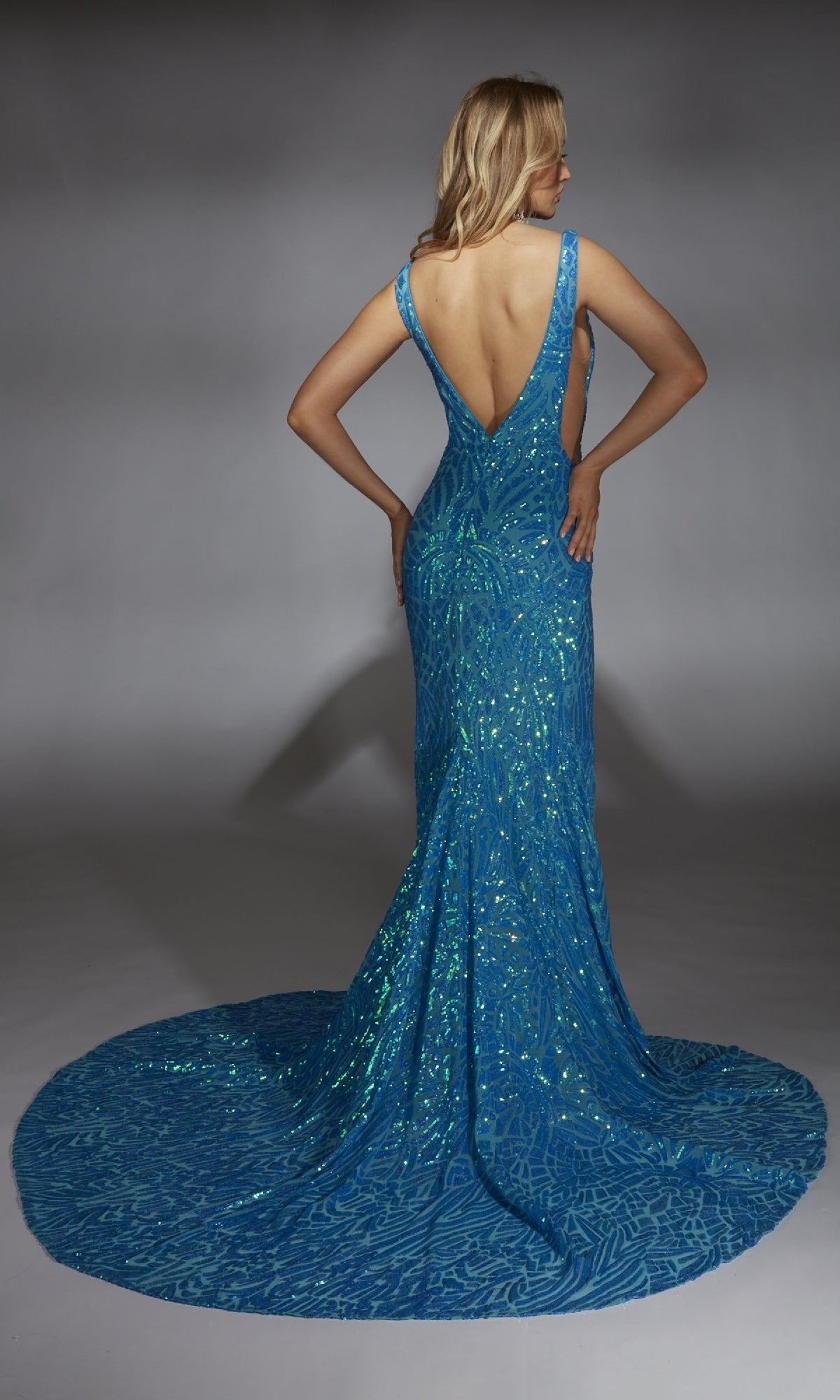 A woman faces away, wearing the Alyce 61876—a form-fitting, floor-length blue sequin prom dress with a deep V-back and flowing train.