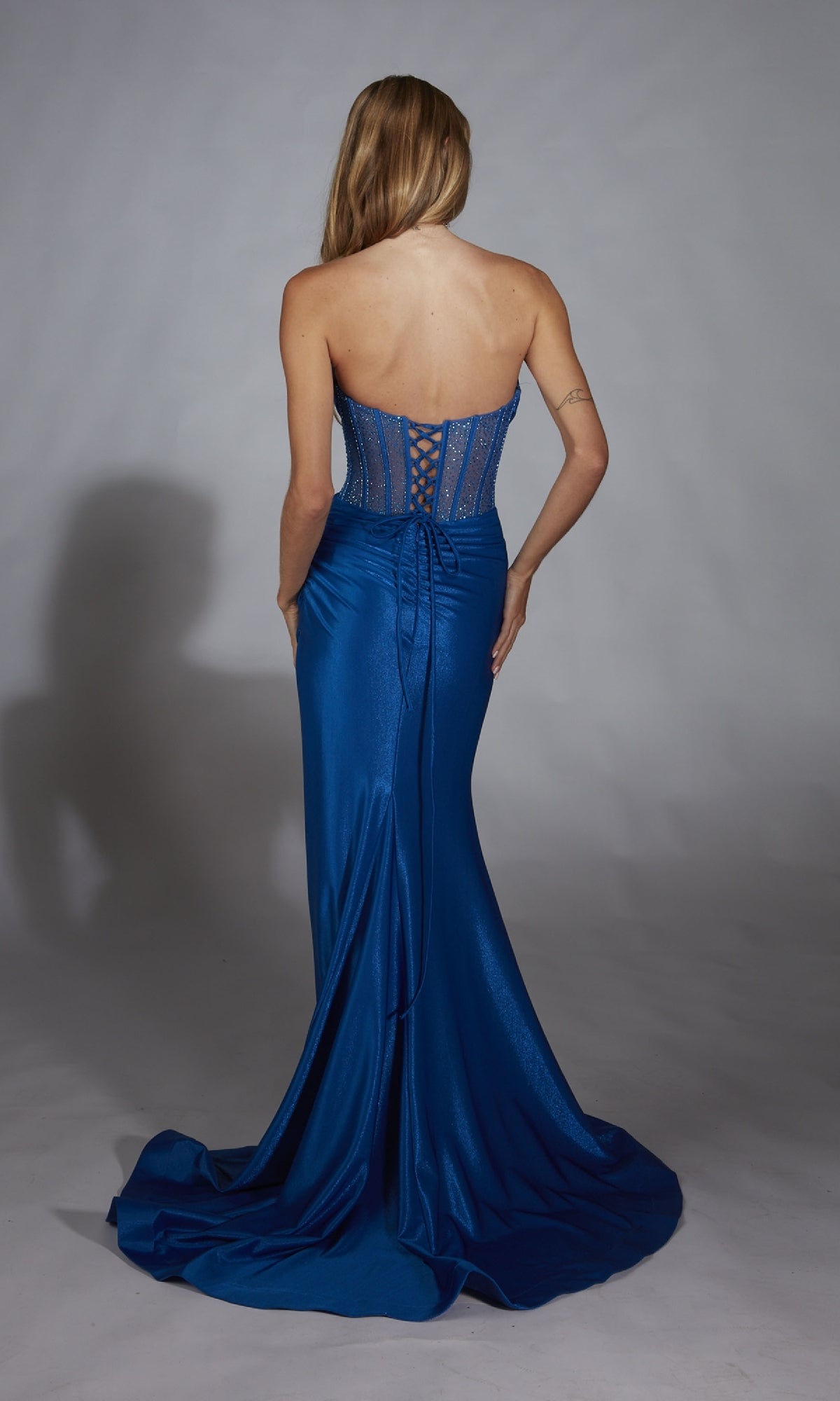 A woman faces away, wearing the Alyce 61882 long prom dress—a strapless, fitted blue gown with a lace-up back and floor-length skirt—set against a plain gray background.