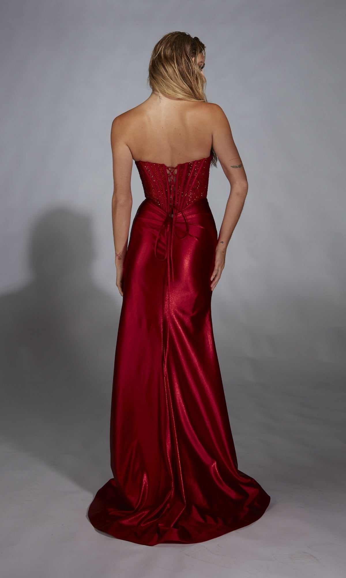 A woman faces away from the camera in the Alyce 61884 long prom dress, a strapless metallic stretch satin gown with a shiny red corset bodice and an eye-catching lace-up back.