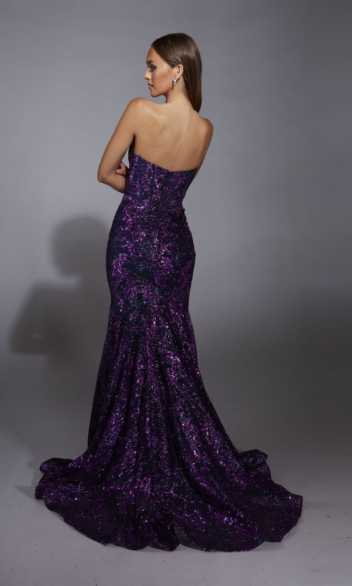 A woman poses with her back to the camera in the Alyce 61887 long prom dress, featuring a strapless mermaid silhouette, shimmering embellishments, and a slight train, set against a plain gray background.